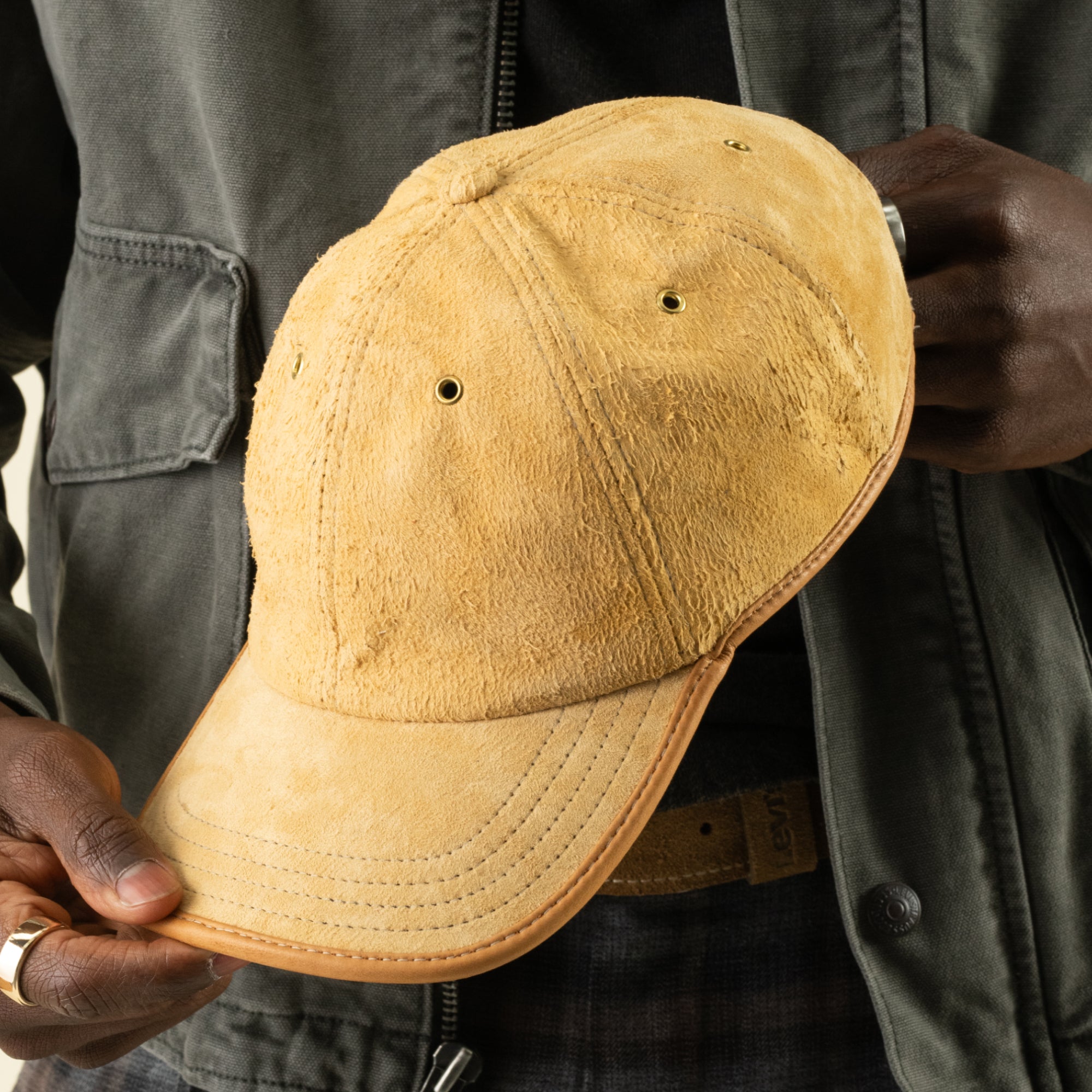 A person in a dark jacket holds the Steiner papa cap by Goorin Bros., featuring tan suede material, visible stitching, ventilation holes, and an adjustable plaid strap at the back.