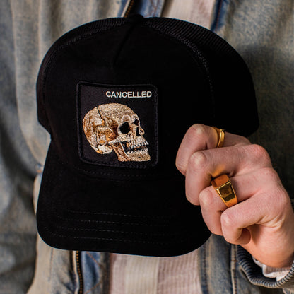 Someone is holding the Bold Suede Skull by Goorin Bros., a limited edition black microsuede trucker hat featuring a stitched skull patch with "CANCELLED" embroidered above it.