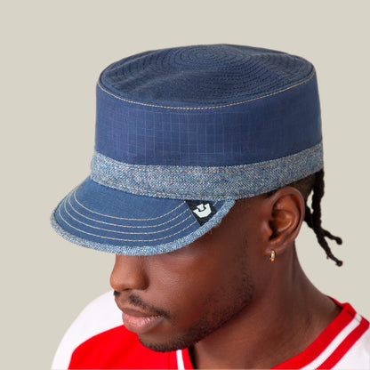 A person wears the Goorin Bros. Ampere, a blue flat-top cadet hat with a denim chambray brim, paired with a red and white shirt, against a plain light background.