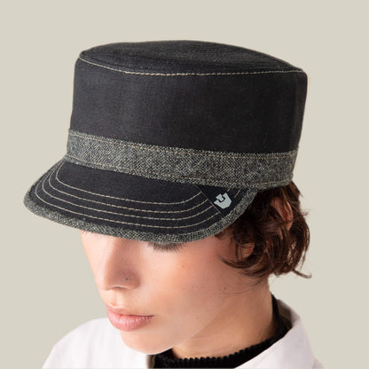A person wearing the Goorin Bros. Ampere flat-top cadet black cap with a grey band and exposed stitching looks downward against a plain light background.