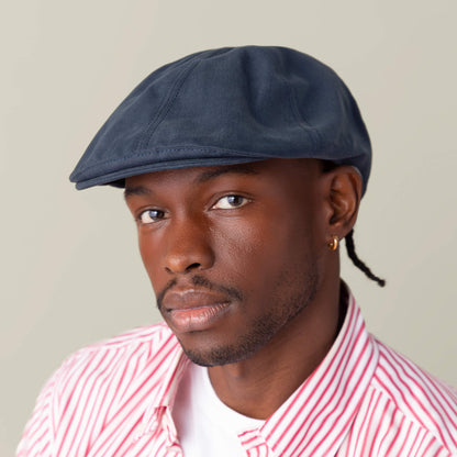 A man wears the Murray Way blue flatcap by Goorin Bros. and a red and white striped 100% cotton shirt over a white t-shirt, looking at the camera against a plain background.