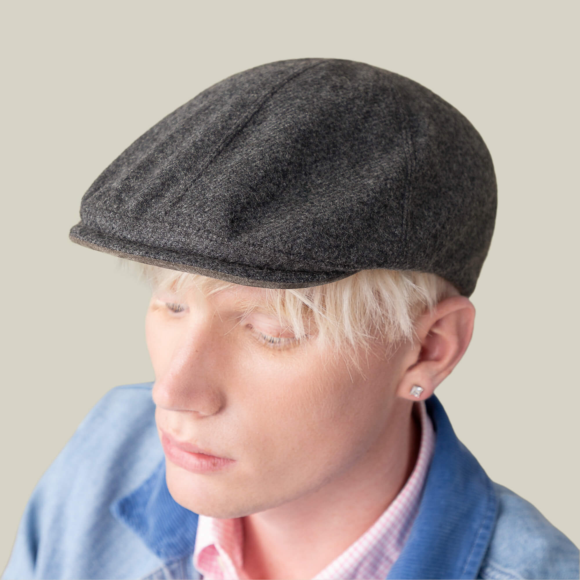 A person with light blond hair wears the Goorin Bros. Ronald Amos dark gray six-panel flat cap, paired with a blue jacket and pink checkered shirt, shown against a plain beige background.