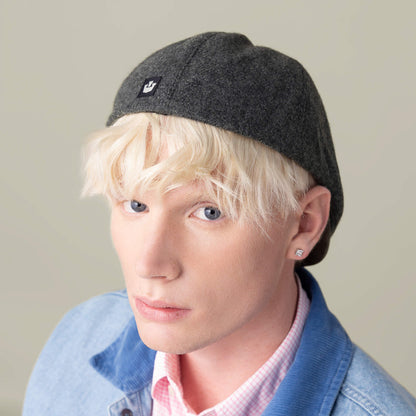 A person with platinum blonde hair wears the Goorin Bros. Ronald Amos gray six-panel hat, blue jacket, and pink checkered shirt, looking at the camera.