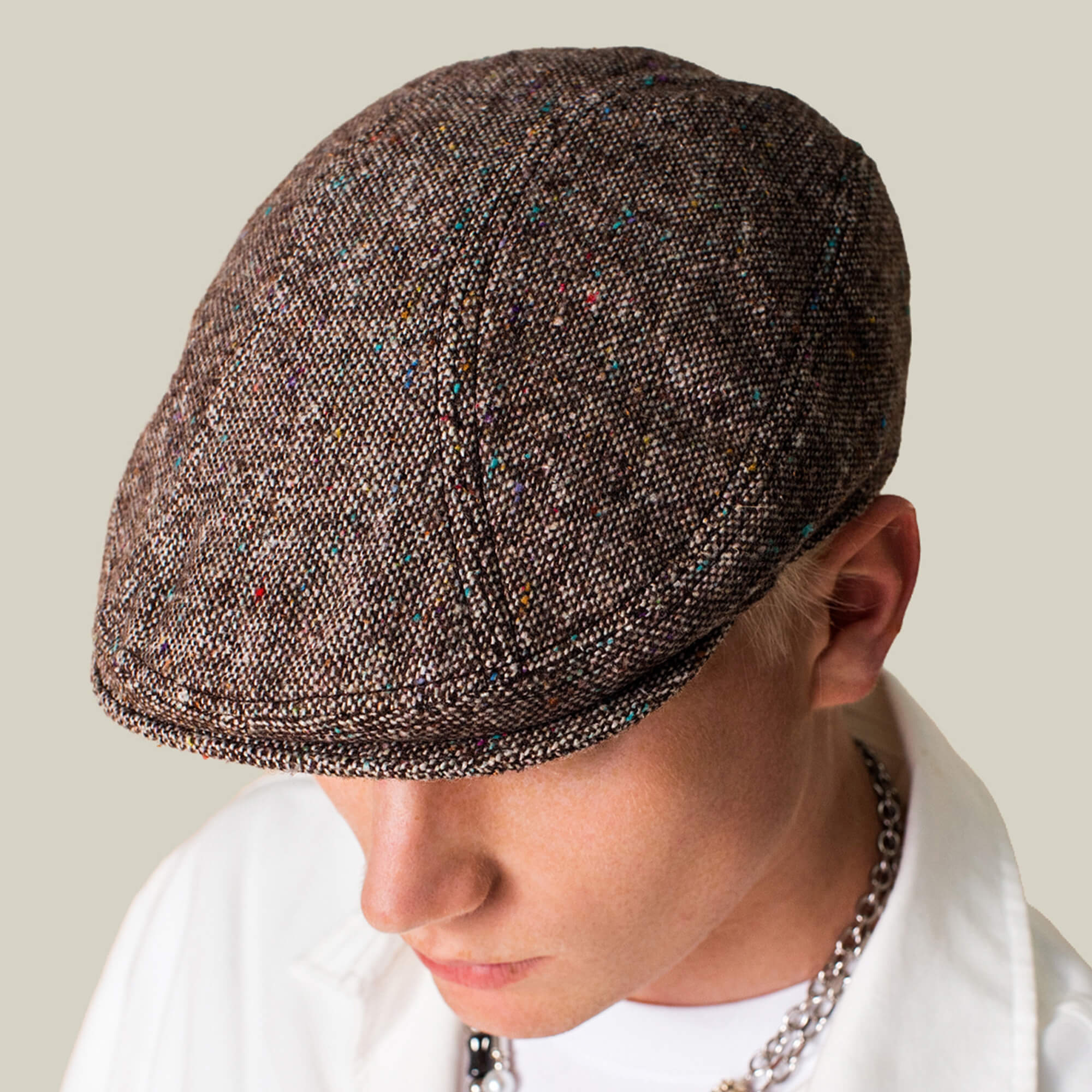 A person wearing the Gibbins grey tweed flatcap by Goorin Bros., a white shirt, and a silver chain necklace against a plain background, highlighting timeless men's style.