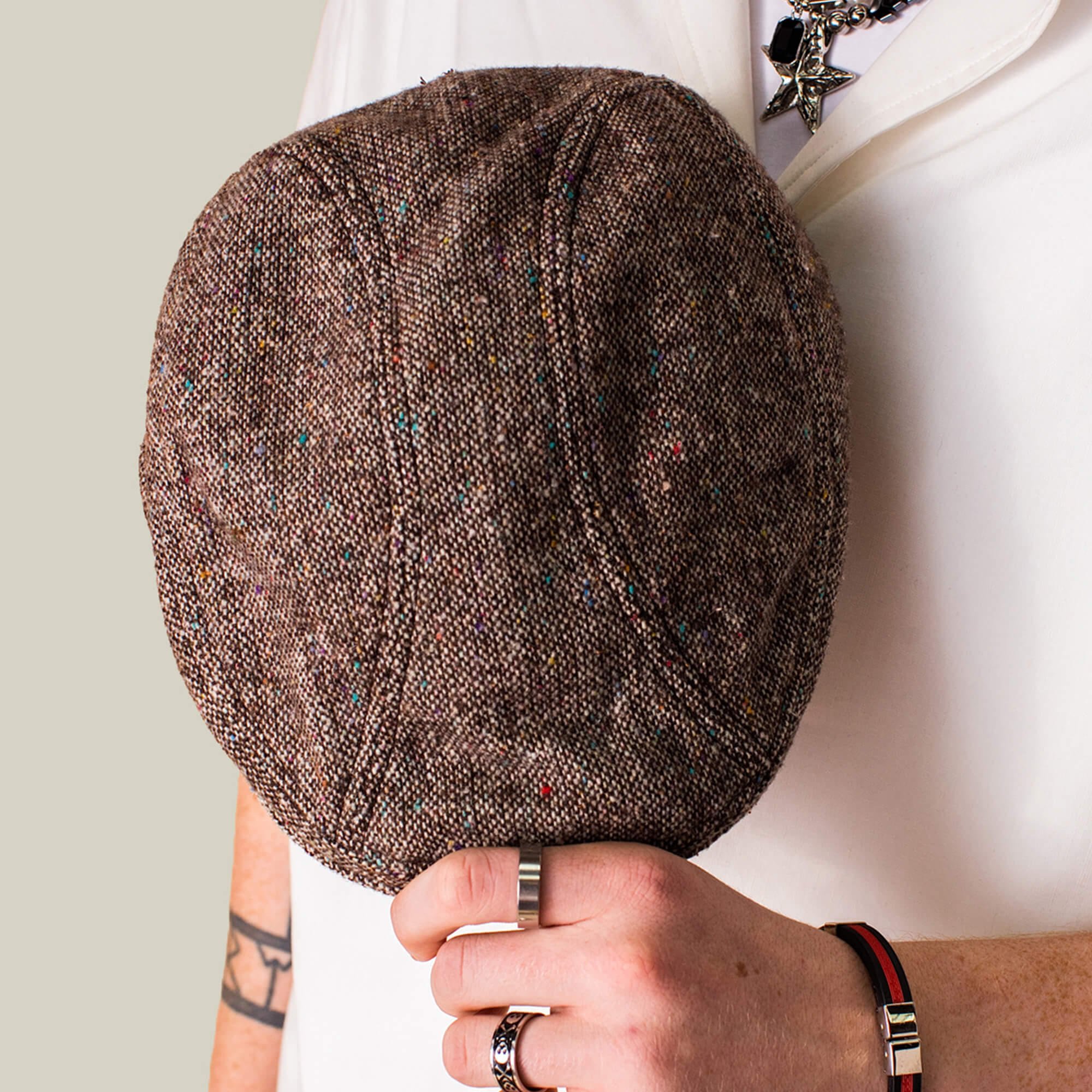 Wearing a white shirt, a person holds the Goorin Bros. Gibbins brown textured flatcap, complemented by rings, a bracelet, and a star pendant necklace for a stylish men's fashion look.
