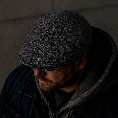 A man in the dim light wears the Goorin Bros. Newlands, a hand cut and sewn grey flecked flat cap, paired with a dark jacket and hoodie, looking downwards.