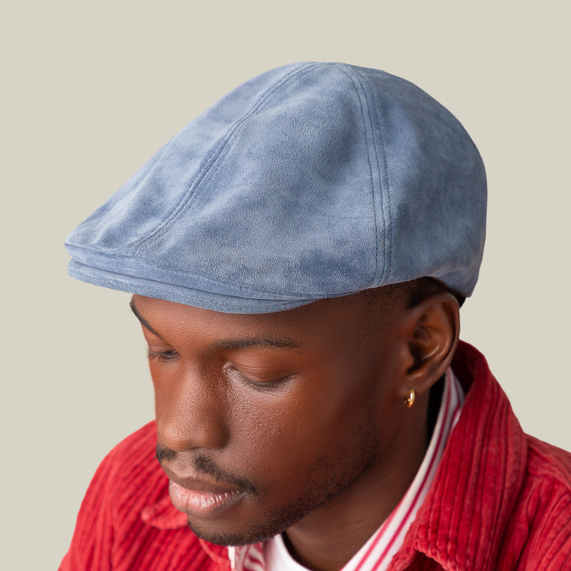 A man in the Cushy Benjamin Paul light blue suede flatcap by Goorin Bros., paired with a red corduroy jacket and striped shirt, looks downward against a plain background.