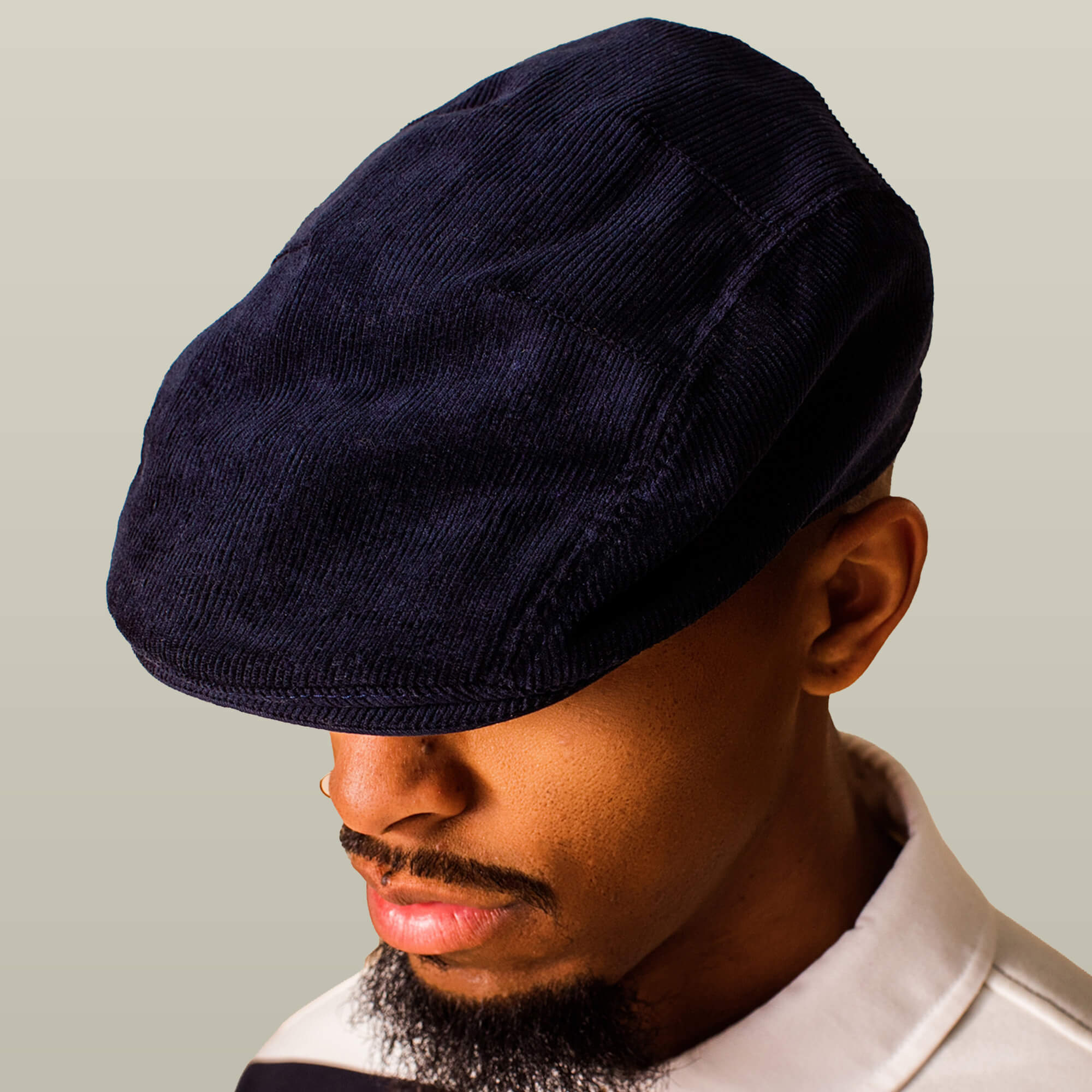 A man wearing the Goorin Bros. Cozy Low Profile, a navy corduroy flatcap, paired with a white collared shirt, faces slightly downward against a plain background.