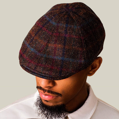 A man in a white collared shirt looks downward, wearing the Cozy Benjamin Paul brown plaid wool duckbill ivy cap by Goorin Bros. against a plain background.