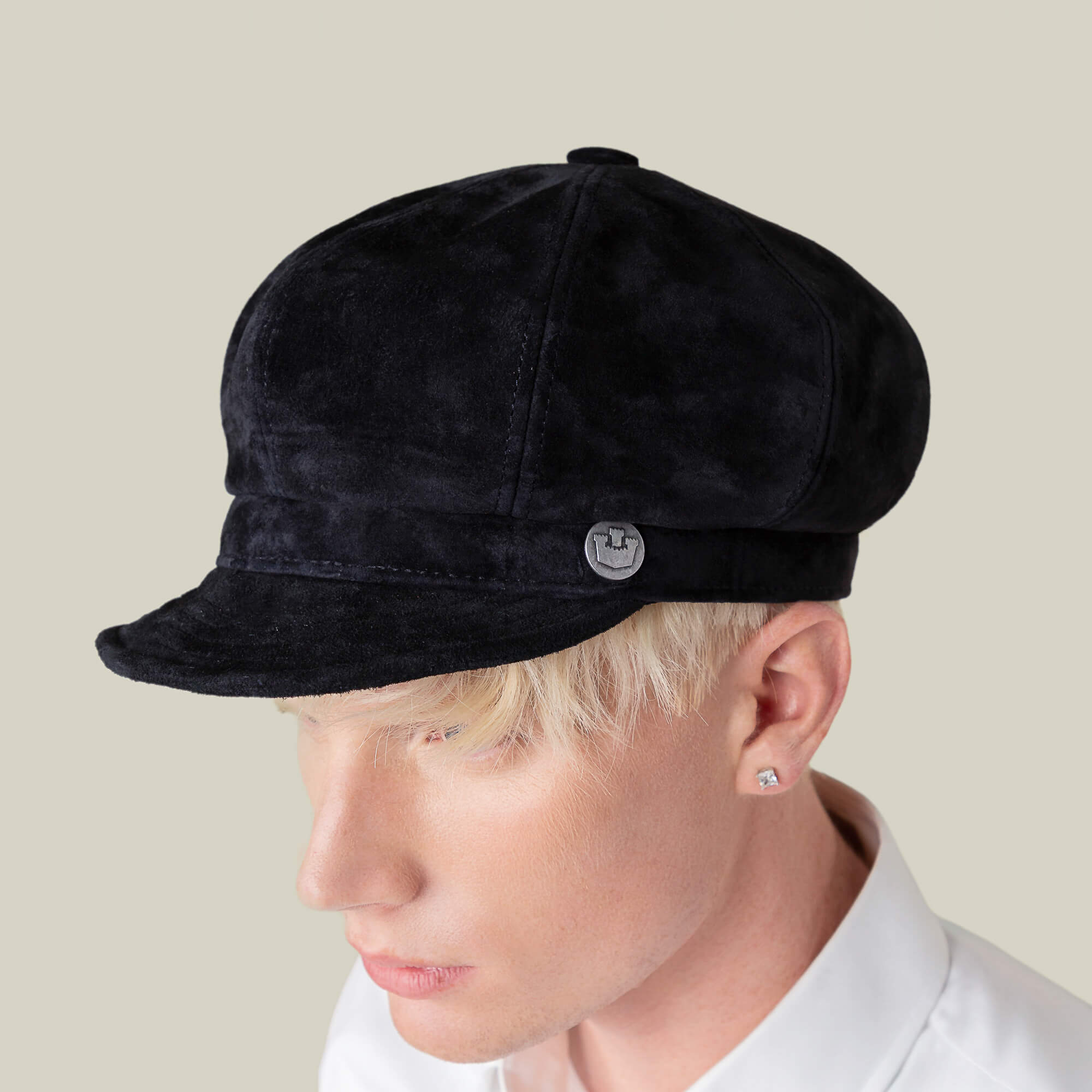 Person wearing the Goorin Bros. Ruthy black suede cabbie flatcap with a metal emblem on the side, styled with a white collared shirt and stud earring against a plain background.