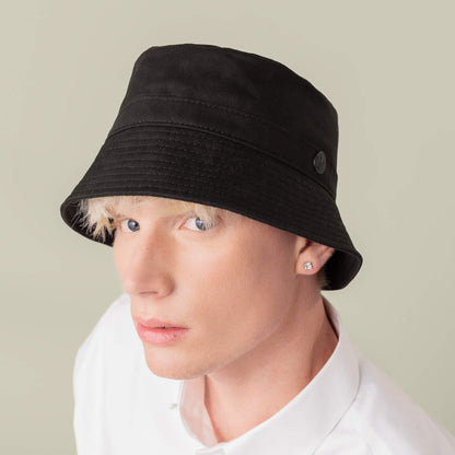Person with light skin and short blond hair wears the Goorin Bros. Odyssey Bucket hat in solid black cotton, paired with a white collared shirt and stud earrings, looking at the camera against a plain background.