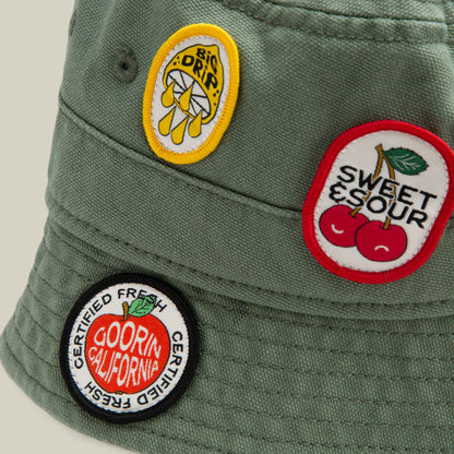 The Goorin Bros. PLU 1895 is an olive green bucket hat souvenir with three colorful embroidered patches: a lemon with BIG DRIP, cherries with SWEET & SOUR, and a round DOORIN CALIFORNIA CERTIFIED FRESH patch.