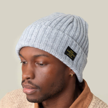A person wearing the Goorin Bros. Dream Cap, a light gray knitted beanie with a black label, along with a beige sweater and brown shirt, is photographed against a plain background.