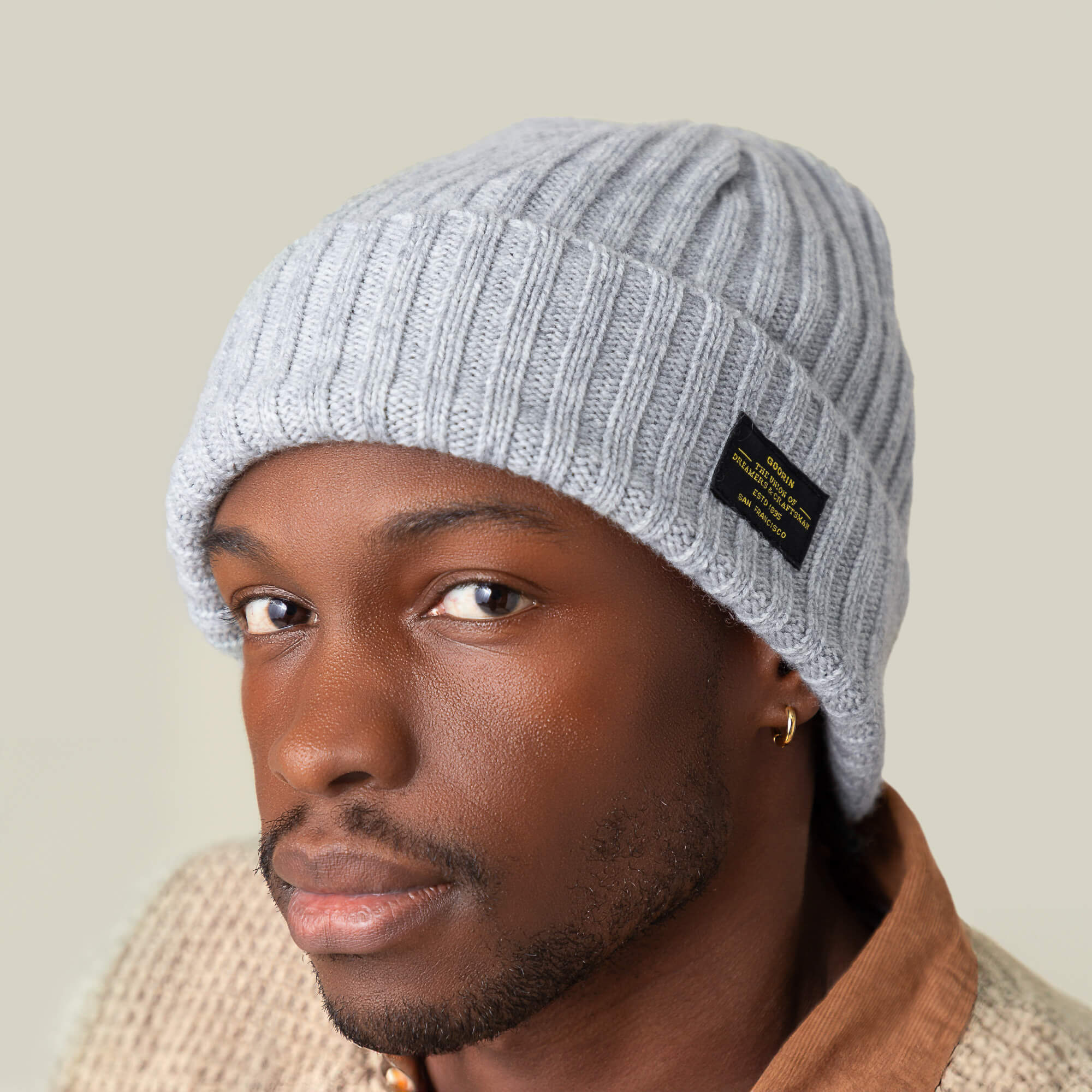 A person wearing the Goorin Bros. Dream Cap, a light gray knitted beanie with a black label, paired with a light brown collared shirt and a luxurious beige textured sweater, looks at the camera.
