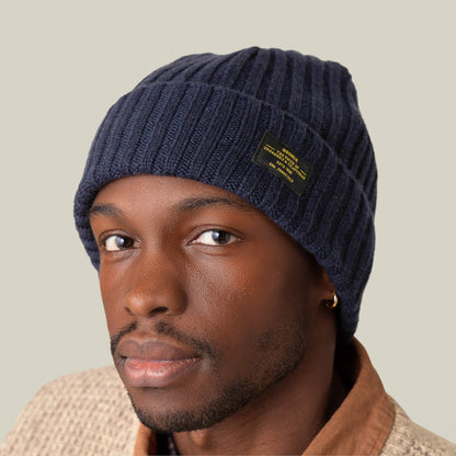 A man wearing the Goorin Bros. Dream Cap, a navy blue cashmere wool beanie with a black label, pairs it with a beige textured jacket and gold hoop earring, gazing at the camera against a plain background.