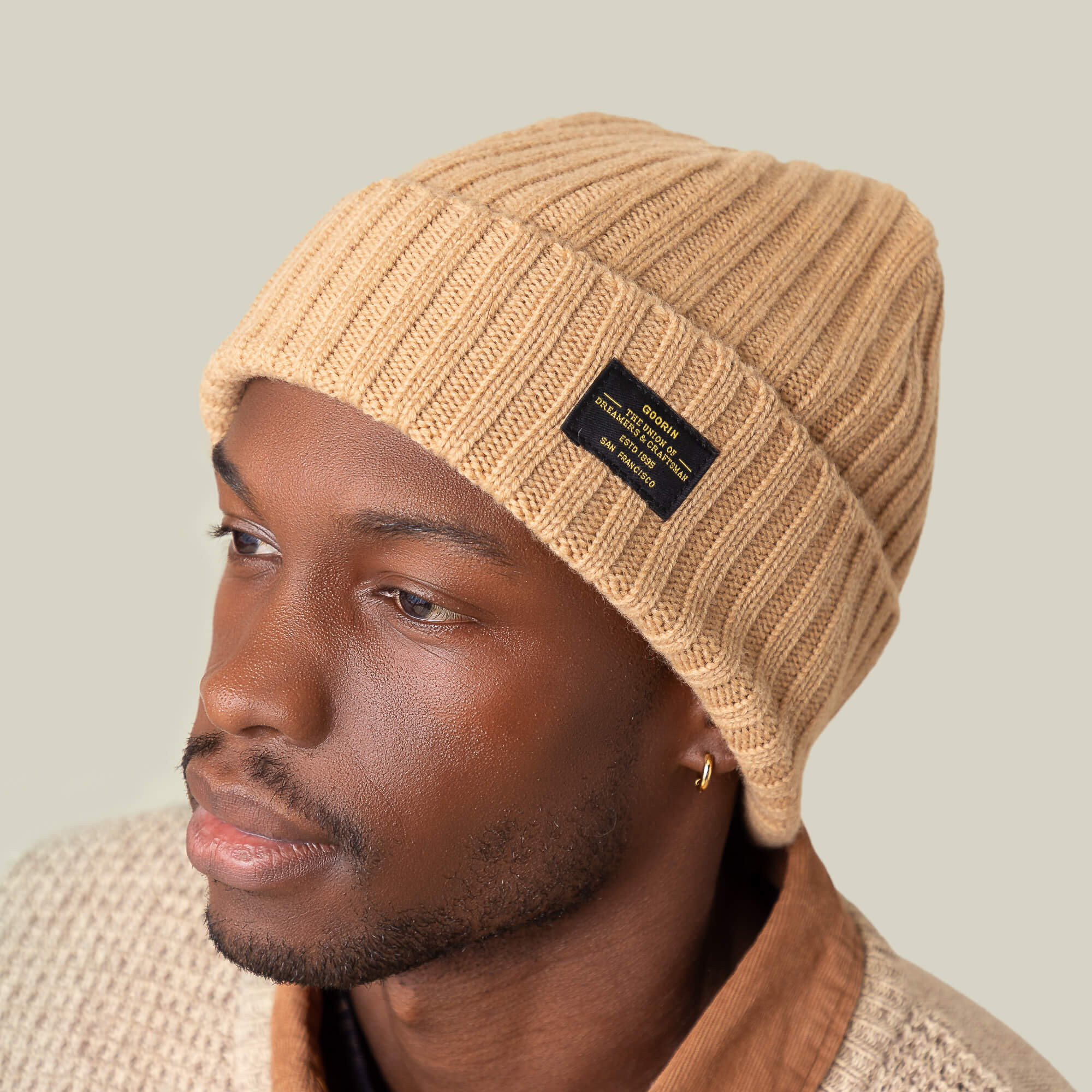 A man wearing the Goorin Bros. Dream Cap, a luxurious beige knitted beanie with a black label, beige textured sweater, and gold hoop earring, looks slightly to the side against a plain background.