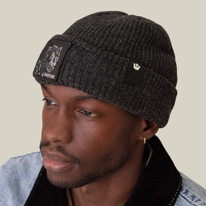 A man wearing The Black Panther Beanie by Goorin Bros.—a dark knit hat featuring a panther patch—paired with a light denim jacket with a black collar, looks slightly to the side against a plain background.