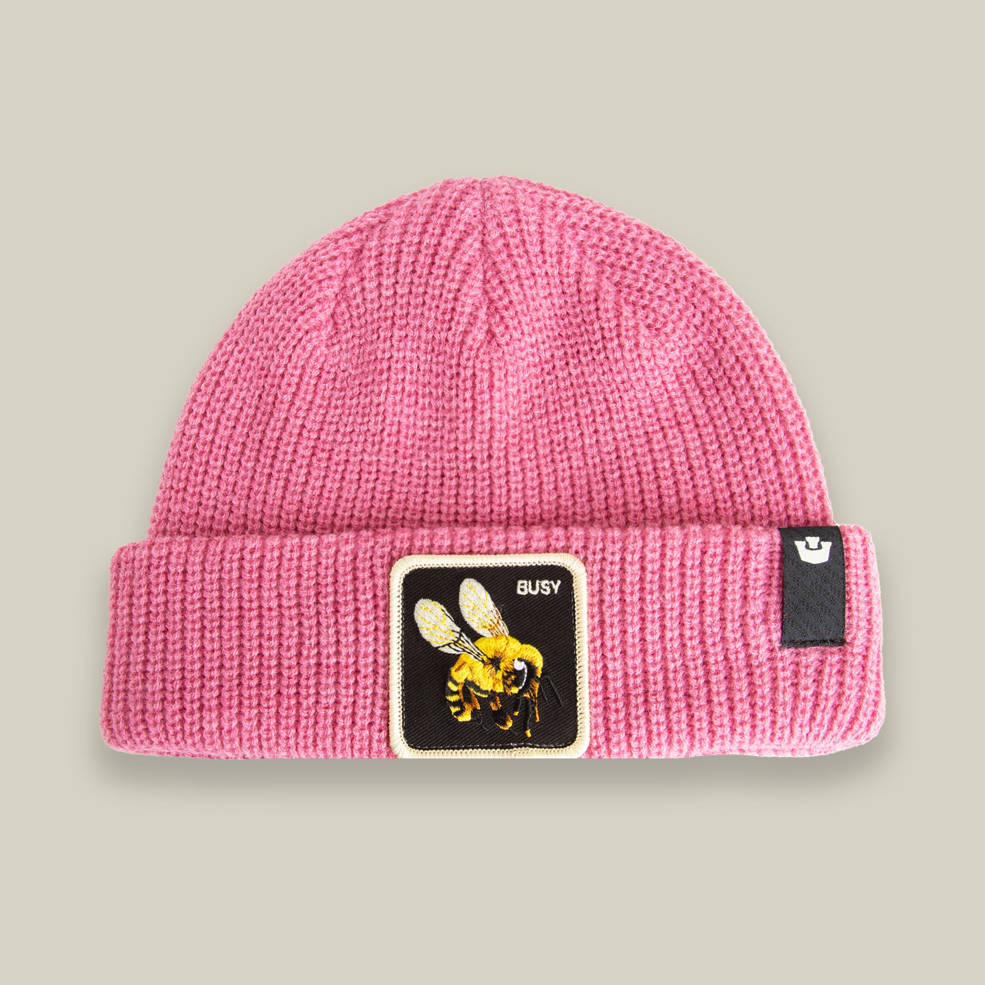 The Busy Beanie by Goorin Bros. is a pink knit hat featuring a buzzing bee patch with "BUSY" and a black tag with a white logo, adding playful warmth to any outfit.
