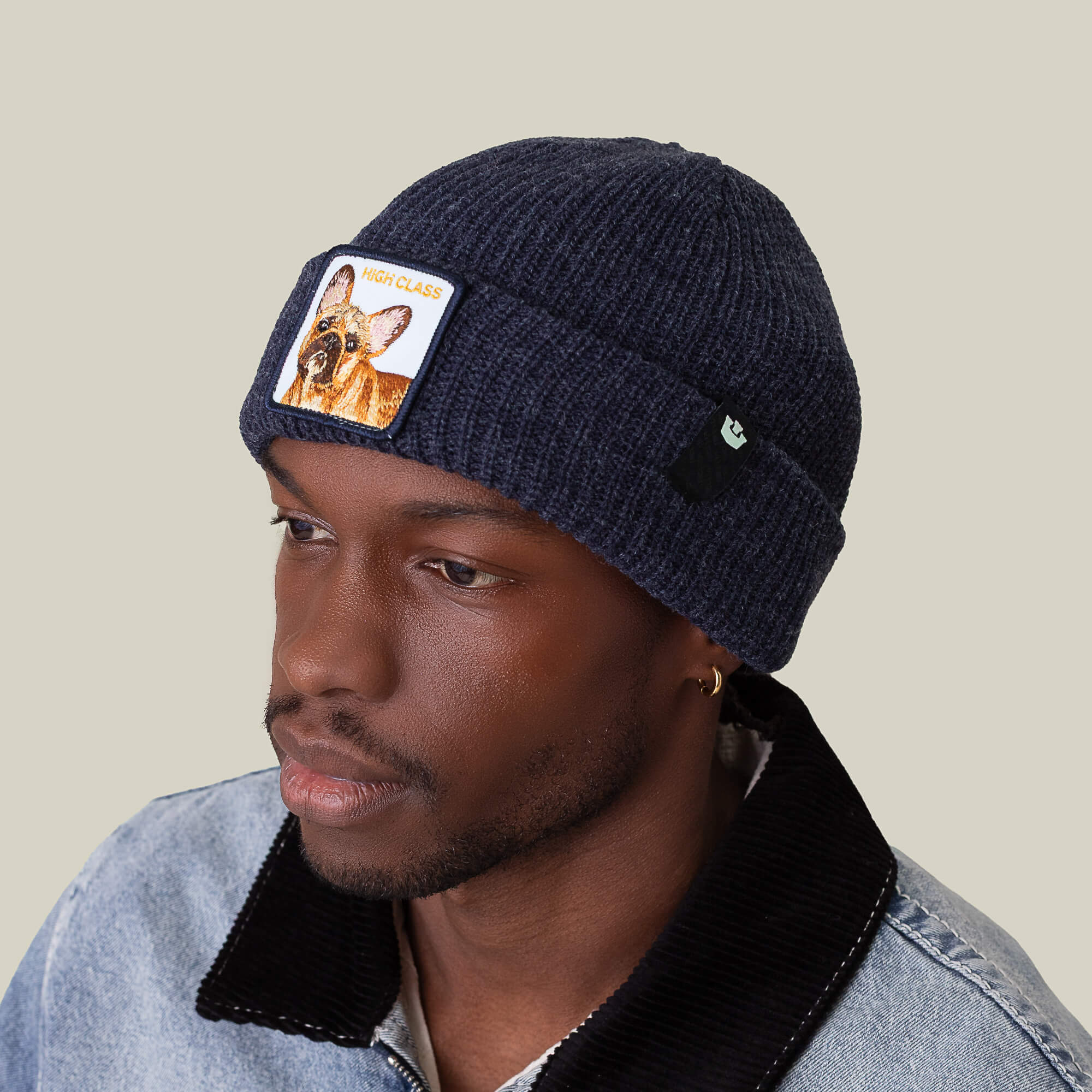 A man wears The High Class Beanie by Goorin Bros.—a navy knit beanie with a dog patch—and a denim jacket with a black collar, looking to the side against a plain background.
