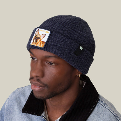 A man wears The High Class Beanie by Goorin Bros.—a navy knit beanie with a dog patch—and a denim jacket with a black collar, looking to the side against a plain background.