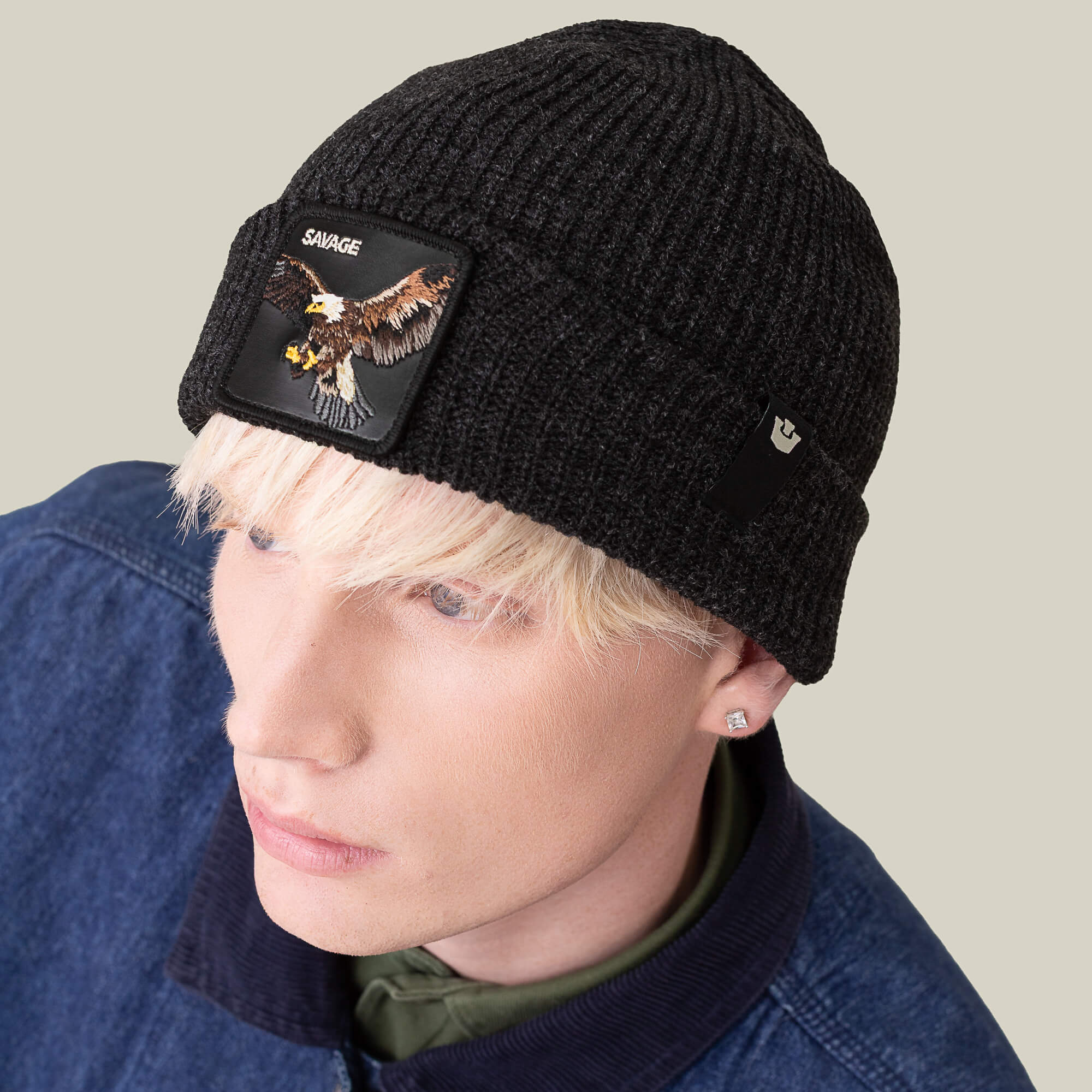 A person with light blond hair wears The Savage Beanie by Goorin Bros.—a black knit hat with an eagle patch labeled SAVAGE—and a dark denim jacket, standing against a plain background.