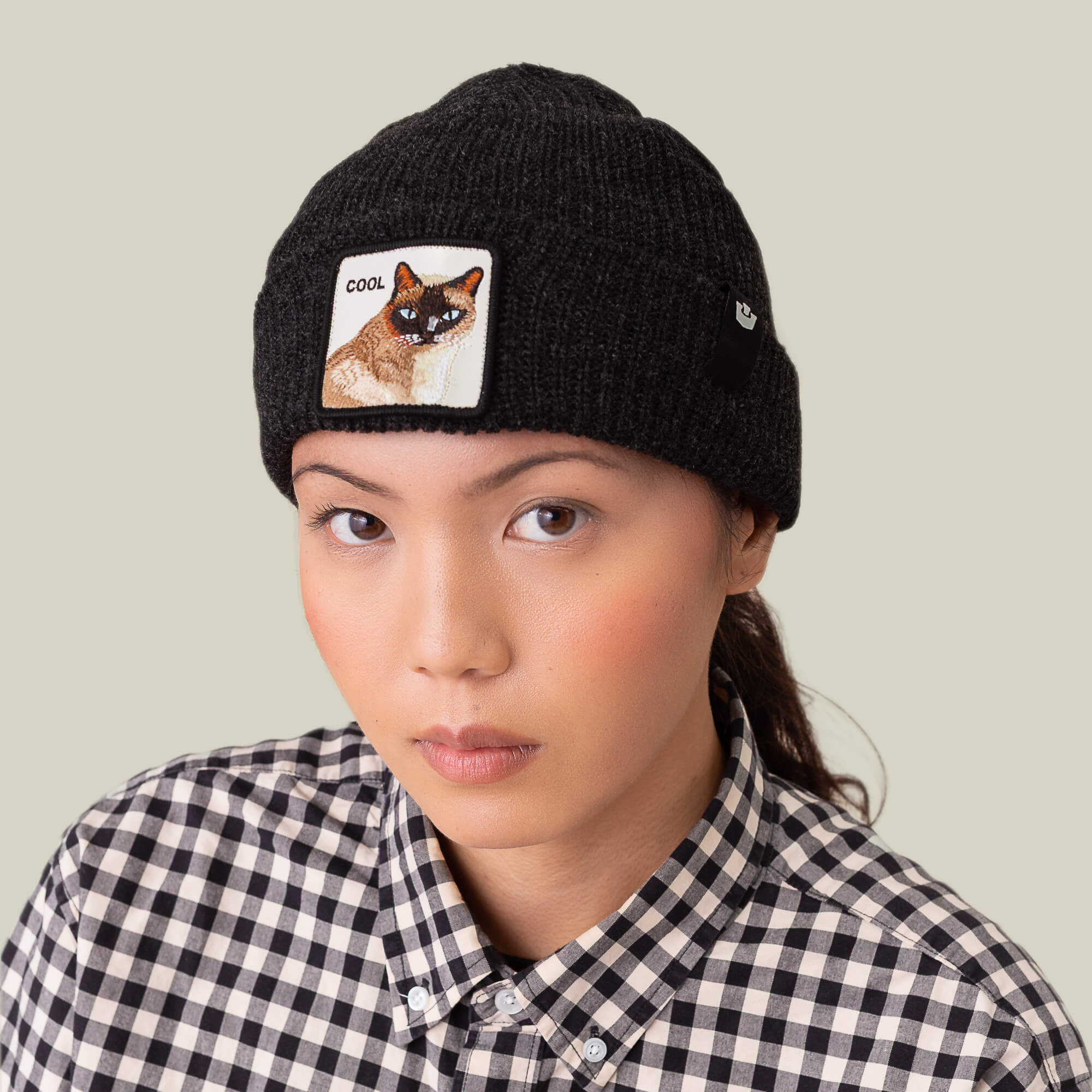 A person wears The Cool Beanie by Goorin Bros., a black ribbed knit hat with a cat patch, paired with a black-and-white checkered shirt, looking at the camera against a plain background.