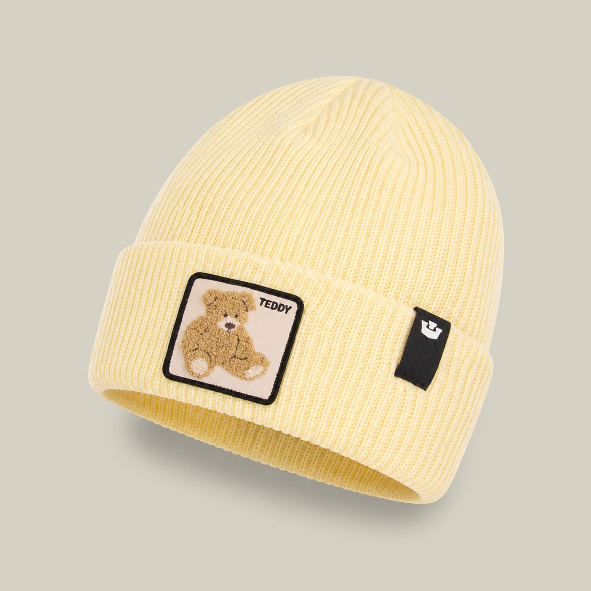 The Double Stuffed - Teddy Beanie by Goorin Bros. is a pale yellow ribbed knit beanie with a folded rib-knit cuff, featuring a teddy bear patch and a small black tag on the front—an adorable winter hat that makes the perfect go-to accessory.