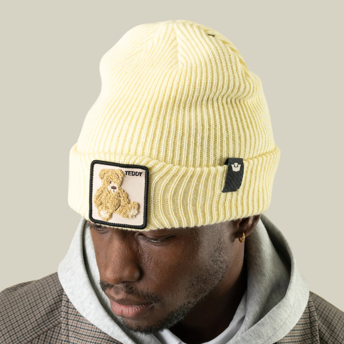 A person wearing a pale yellow rib-knit Double Stuffed - Teddy Beanie with a square patch featuring a teddy bear and the word TEDDY.