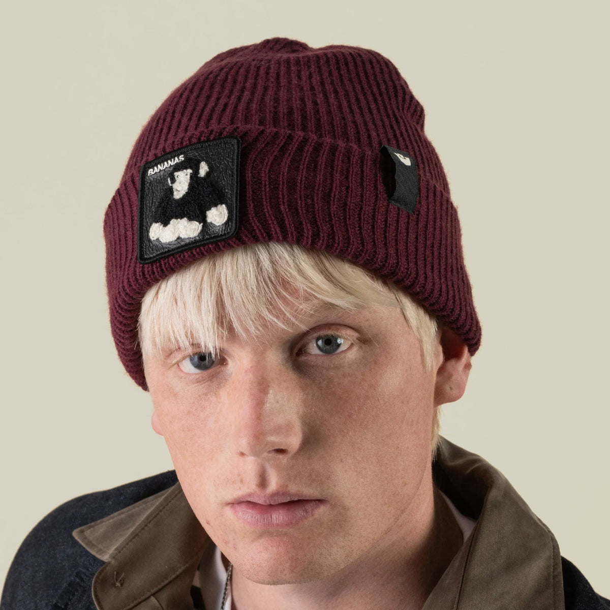 A person wearing a maroon rib knit Double Stuffed - Bananas Beanie featuring a patch of a gorilla and the word BANANAS.