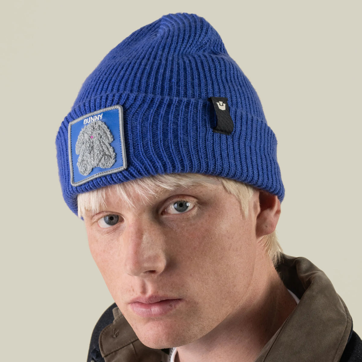 A person wearing a blue rib-knit cuffed Double Stuffed - Bunny Beanie featuring a patch of a gray rabbit and the word BUNNY.