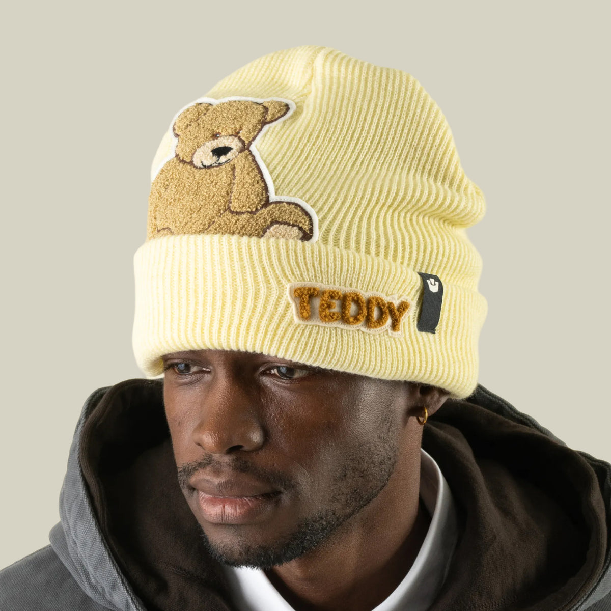 A person wearing a pale yellow, oversized, ribbed Double Stuffed - Teddy Beanie - big featuring a teddy bear patch and the word TEDDY embroidered on the cuff.