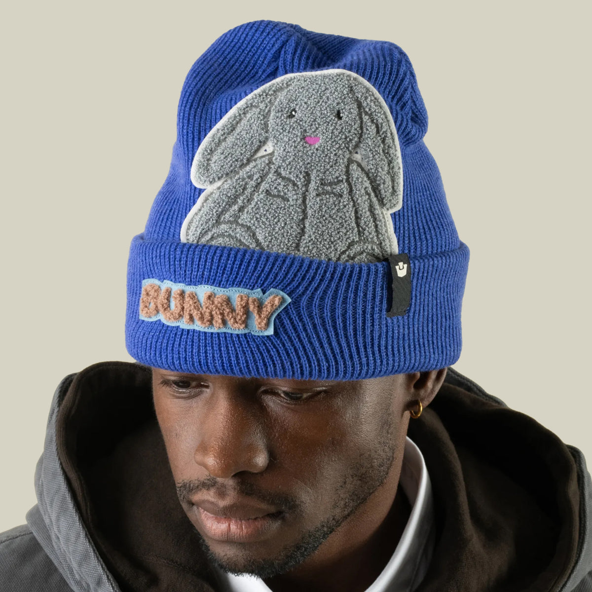 A person wearing a blue Double Stuffed - Bunny Beanie - big, featuring a plush gray bunny patch on the crown and a textured patch on the folded cuff.