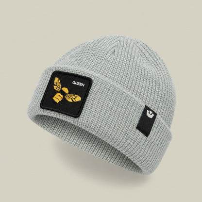 The Queen Bee Beanie by Goorin Bros is a light gray ribbed knit beanie featuring a black embroidered patch with a gold queen bee and "QUEEN" text on the front, displayed on a neutral beige background.