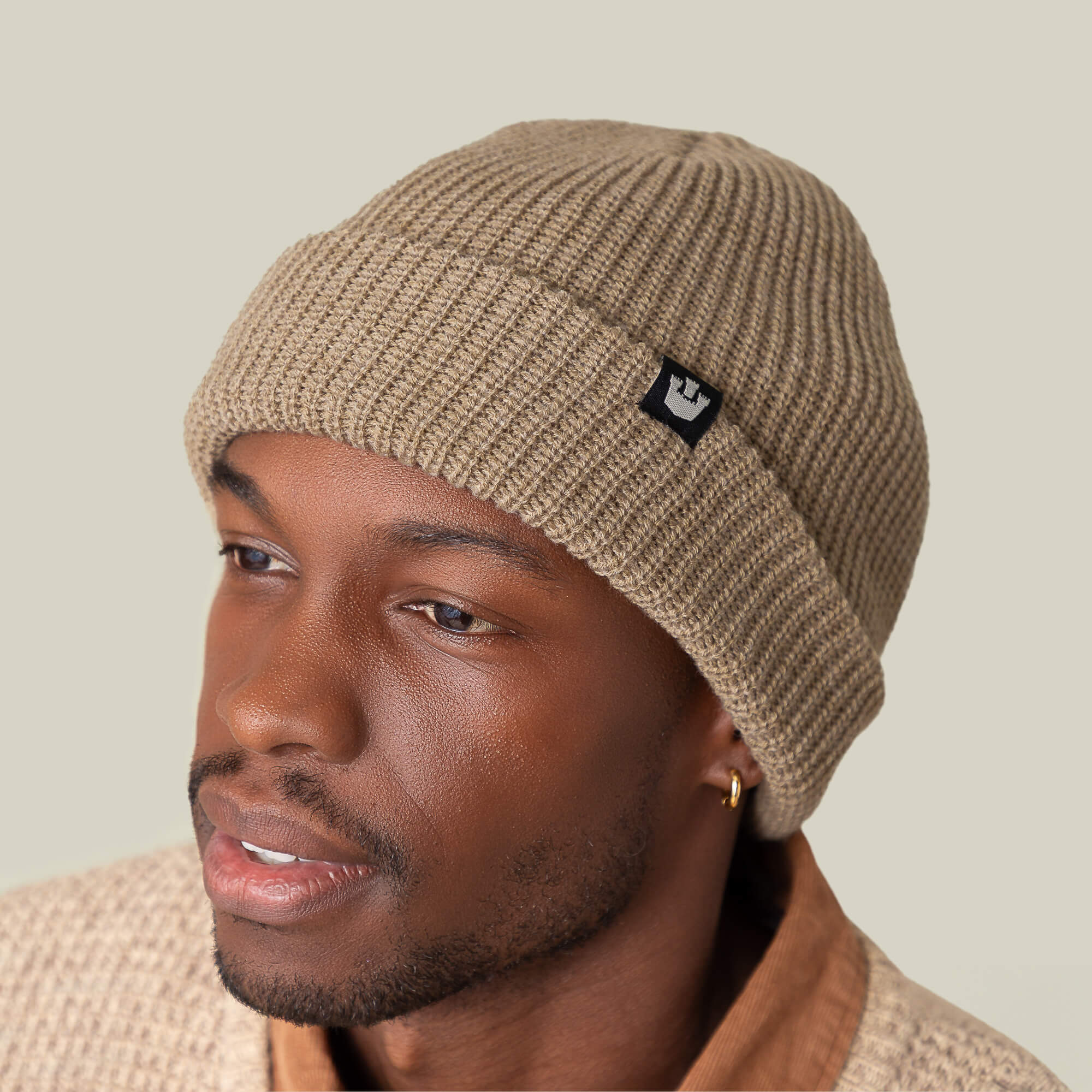 Someone wears the Goorin Bros. Air to Fakie beige Watchman beanie with a black logo and a matching textured sweater, glancing sideways against a plain background. Its lightweight poly knit makes it ideal for all-season use.