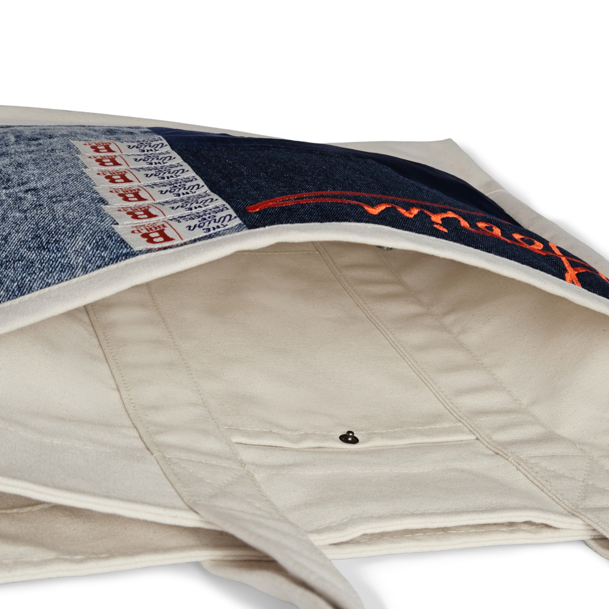 A close-up of the Goorin Bros. Goorin Reworked Tote highlights its interior lining, snap button closure, and a blue denim patch with red stitching and woven labels.