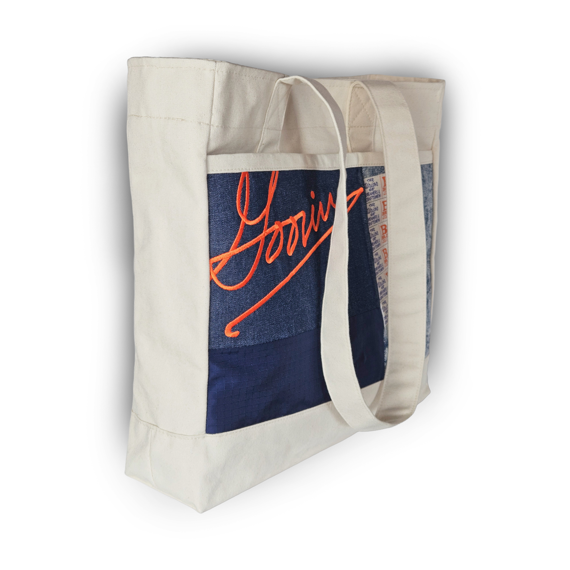 The Goorin Bros. Goorin Reworked Tote is a white canvas bag with long handles, a blue patch, and orange “Goorin” script. Made in the USA from upcycled fabric for an eco-friendly, stylish look.