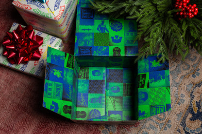 An open, empty Goorin Gift Box by Goorin Bros. with a blue and green interior sits on a patterned surface beside a wrapped present and festive pine branches—an ideal gift for any headwear lover or Goorin Bros. fan.