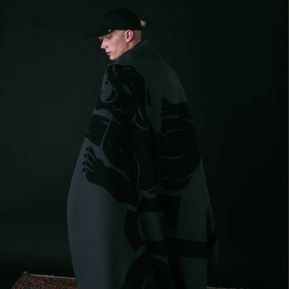 A person in a black cap stands against a dark background, wrapped in the Goorin Bros. Panther Crest Blanket featuring a bold animal graphic.
