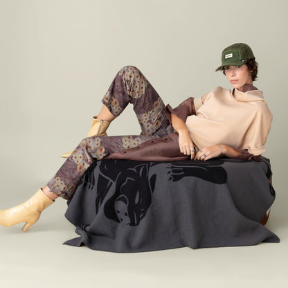 A person in a beige top, patterned pants, yellow boots, and green cap lounges on a seat draped with the Goorin Bros. Panther Crest Blanket, which showcases a bold panther design.