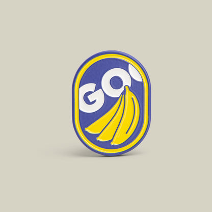 The Goorin Bros. GO Bananas Pin in ROYAL features "GO!" in white above three yellow bananas on a blue and yellow oval background—a playful accessory for any outfit.