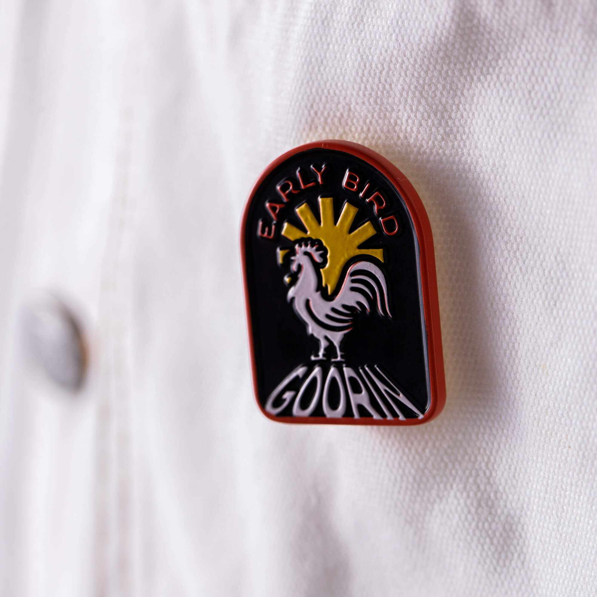 The Early Bird Pin by Goorin Bros., featuring a rooster and sun rays with "EARLY BIRD GOORIN" text, is shown attached to white fabric.