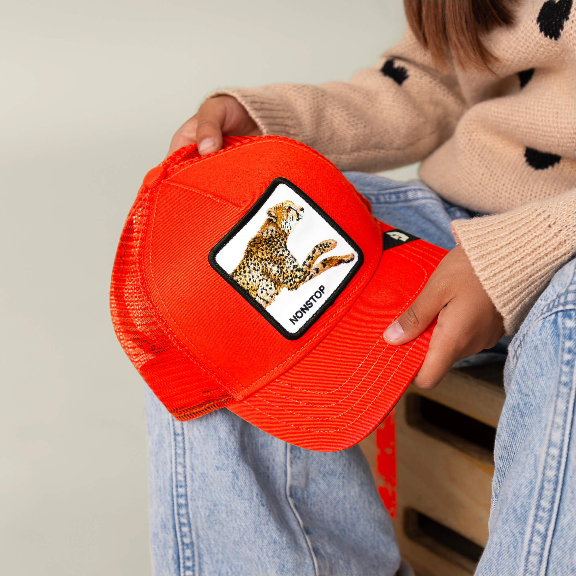 A person in jeans and a beige sweater with black dots holds The Nonstop Cheetah Mini cap by Goorin Bros., an orange kids cap featuring a cheetah image and the word "NONSTOP" on the front patch.