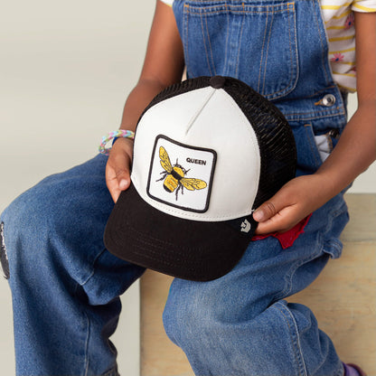 A child in denim overalls holds Goorin Bros.’ The Queen Bee Mini, a black-and-white kids cap featuring an embroidered crowned bee and the word QUEEN.
