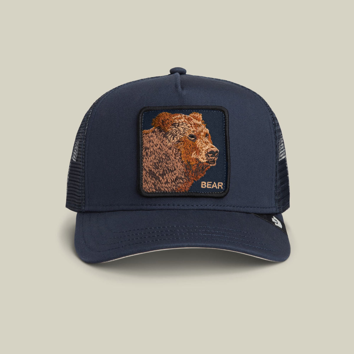The Goorin Bros. Bear Trucker Mini is a navy blue trucker hat with a mesh back, featuring an embroidered brown bear patch with the word "Bear" on the front panel, displayed against a neutral beige background.