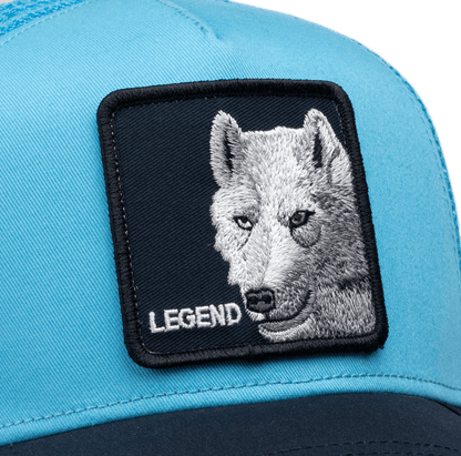 The Goorin Bros. Legend Wolf Trucker Mini is a light blue and black two-tone snapback cap featuring an embroidered white wolf head patch with yellow eyes and the word "Legend" on a dark navy background.