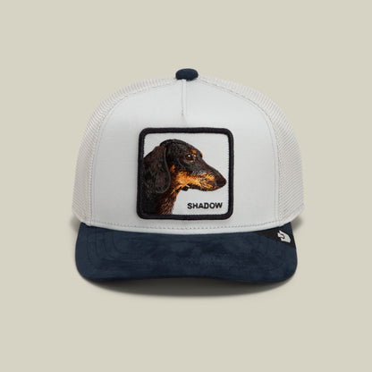 The Goorin Bros. Suede Shadow Mini is a white and navy trucker hat featuring a mesh back and embroidered dachshund patch with text reading "Shadow" on the front.