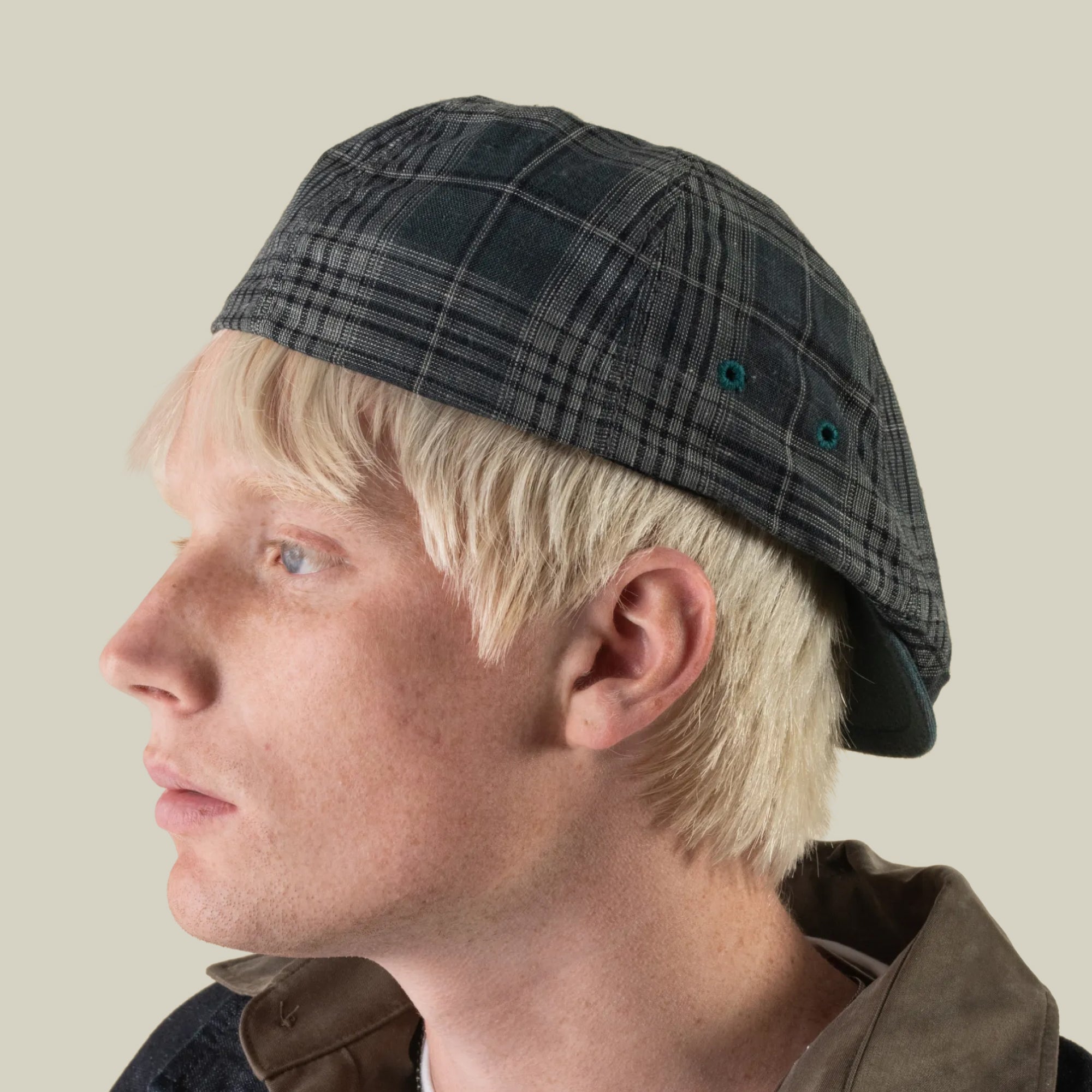A person with light blonde hair wears the Point Lobos 6 Panel Ivy hat by Goorin Bros., a dark plaid cap crafted from a breathable cotton blend and handmade in the USA, paired with a brown collared jacket, facing left against a plain light background.