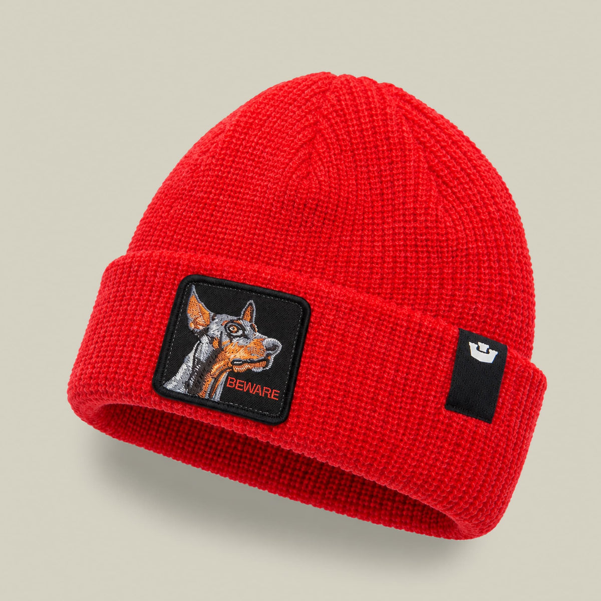 The Beware Beanie Mini by Goorin Bros is a bright red ribbed knit beanie featuring a black patch with an embroidered dog and "BEWARE" text on the front, displayed against a neutral background.