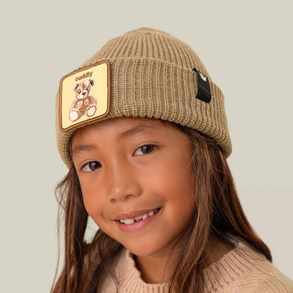 A smiling child wears The Cuddly Beanie Mini by Goorin Bros., featuring a teddy patch, paired with a beige sweater against a plain background.