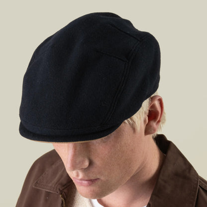A person wearing the Goorin Bros. Cornerstone dark wool flat cap and a brown jacket, facing downward against a plain light background.
